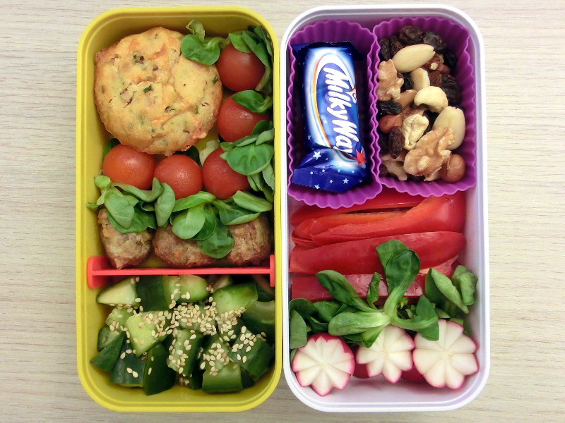 Bentobox gefüllt mit Käse-Schinken-Muffin, Tomaten, Hackbällchen, Gurke, Schokolade, Studentenfutter, Paprika und Radieschen.