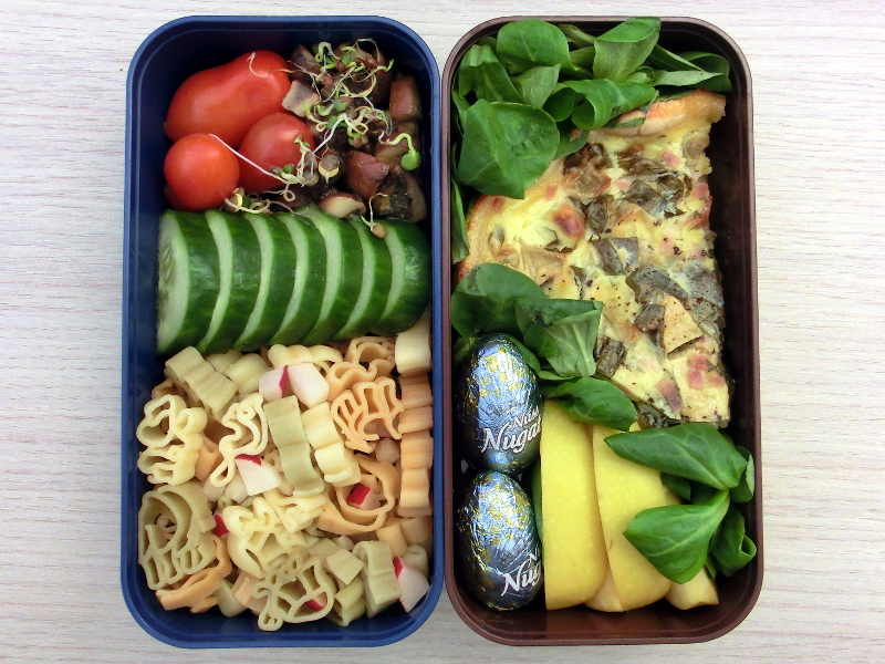 Bento Box gefüllt mit Tomaten, Gurke, Pilzpfanne, Quiche, Nodelsalat, Apfel, Schokolade