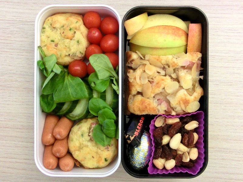 Bento Box gefüllt mit Muffin, Tomaten, Gurke, Würstchen, Apfel, Rhabarberkuchen, Studentenfutter, Schokolade