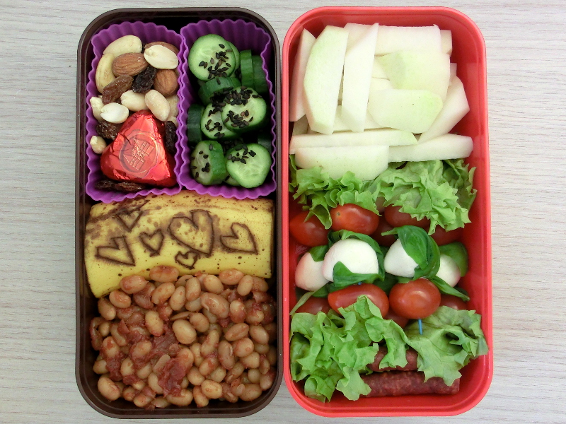 Bento Box gefüllt mit Kohlrabi, Tomate-Mozzarella Spieße, Würstchen, Backed Beans, Banane, Schokolade, Studentenfutter, Gurke