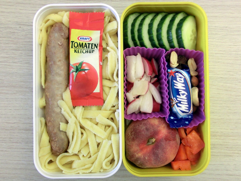Bento Box gefüllt mit Nudeln mit Ketchup, Bratwurst, Gurke, Radieschen, Studentenfutter, Schokolade, Pfirsich, Paprika
