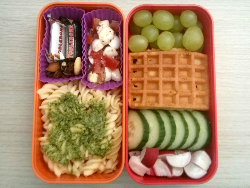 Bento Box gefüllt mit Gurke, Radieschen, Waffeln, Weintrauben, Tomate-Mozzarella, Toblerone, Studentenfutter, Nudeln mit Pesto