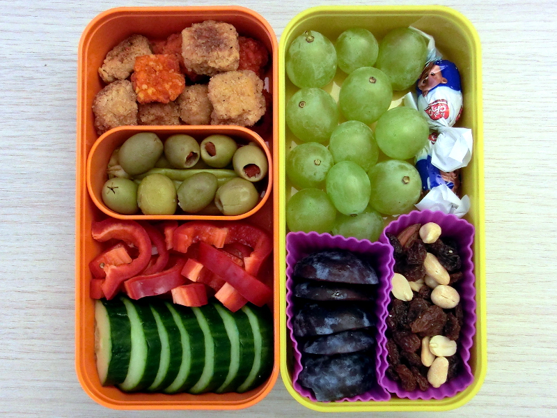 Bento Box gefüllt mit Gurke, Paprika, Oliven, Hähnchen-Nuggets, Peperonie, Weintrauben, Schokolade, Pflaumen, Studentenfutter