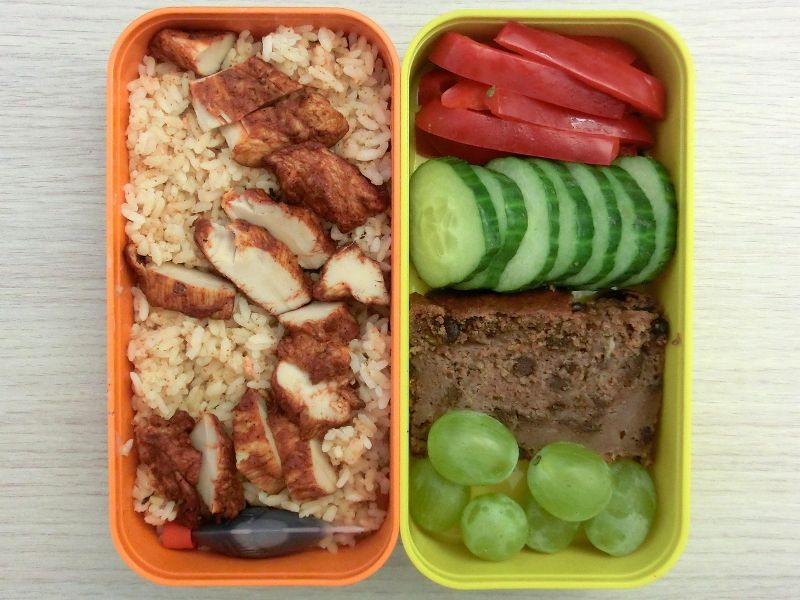 Bento Box gefüllt mit Reis mit Hühnchen, Weintrauben, Bananen-Schokokuchen, Gurke, Paprika
