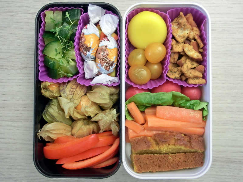 Bento Box gefüllt mit Gurke, Schokolade, Physalis, Paprika, Tomaten, Babybel, Pretzels, Radieschen, Karotten, Kürbisbrot