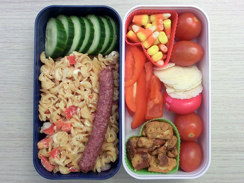 Bento Box gefüllt mit Linsennudeln, Salami, Gurke, Pretzels, Tomaten, Babybel, Parmesancracker, Paprika, Amerikanisches Zuckerzeug