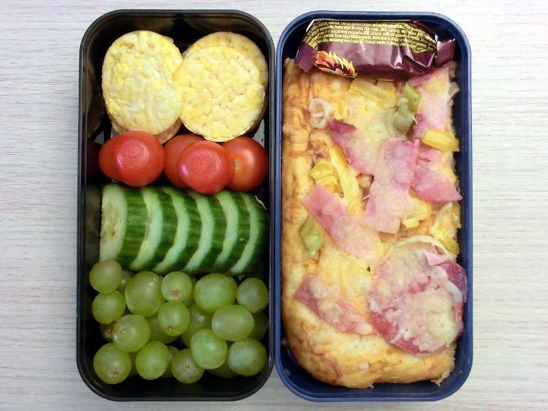 Bento Box gefüllt mit Pizza, Schokolade, Maiscracker, Tomaten, Gurke, Weintrauben