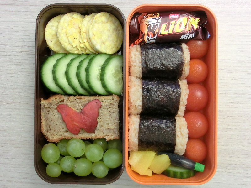 Bento Box gefüllt mit Paprika, Gurke, Tomaten, Onigiri, Schokolade, Maiscracker, Bananenkuchen, Weintrauben