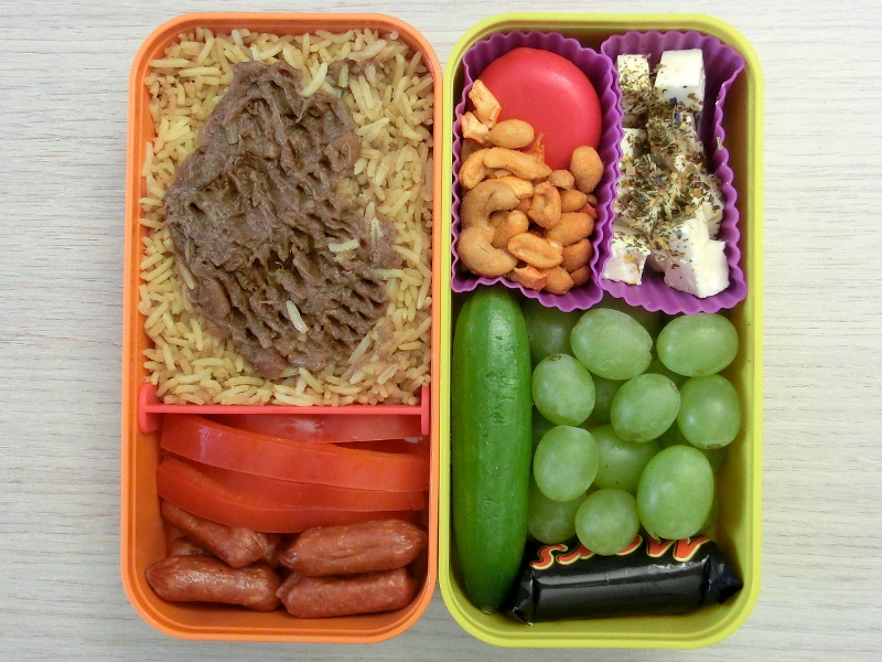 Bento Box gefüllt mit Reis mit Chutney, Paprika, Cabanossi, Babybel, Nüsse, Schafskäse, Weintrauben, Gurke, Schokolade