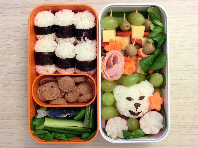 Bento Box gefüllt mit Onigiri, Kekse, Gurke, Käse, Paprika, Weintrauben, Radieschen
