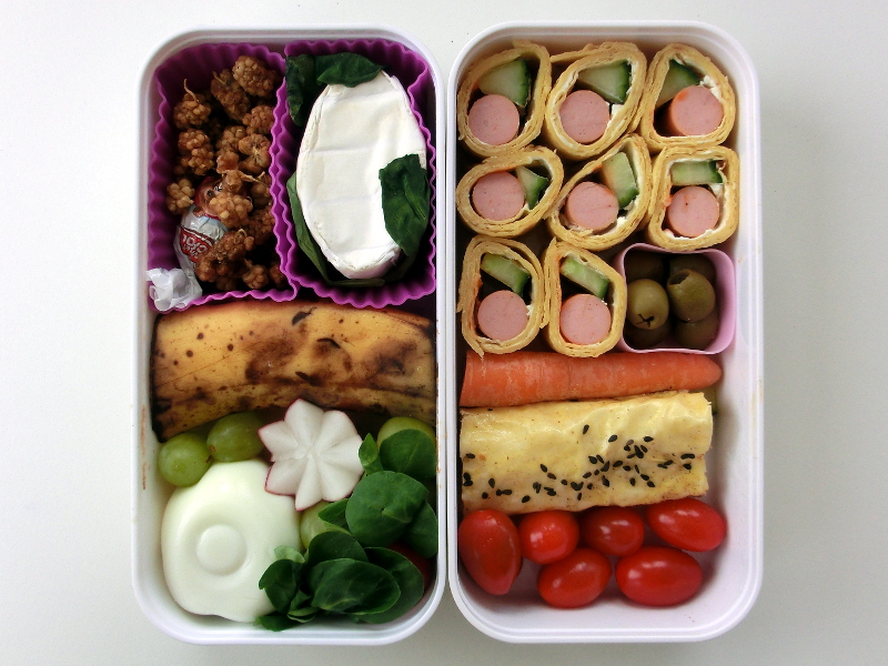 Bento Box gefüllt mit Käse, Banane, Ei, Radieschen, Wraps, Würstchen, Oliven, Möhren, Börek, Tomaten, Schokolade