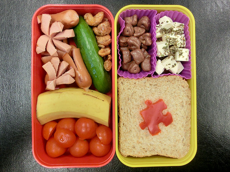 Bento Box gefüllt mit Sandwich, Käse, Schokolade, Erdnüsse, Gurke, Würstchen, Banane, Tomaten