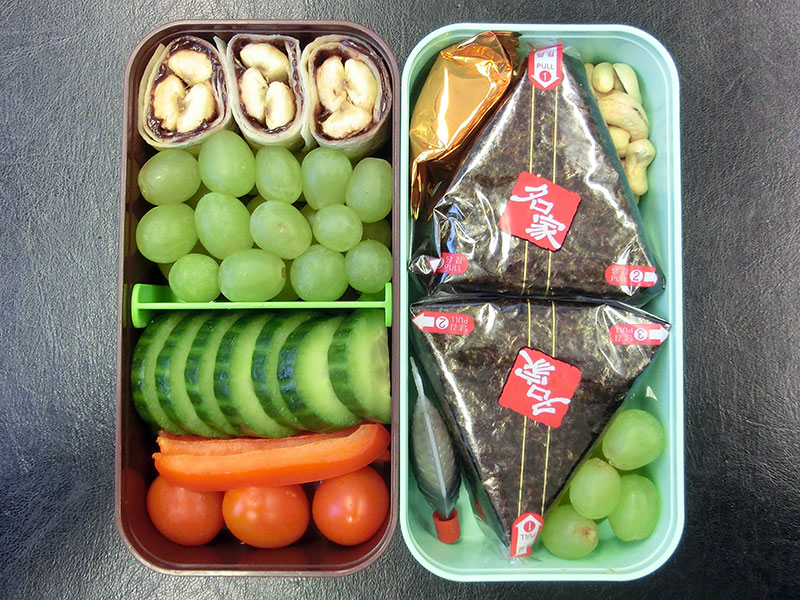 Bento Box gefüllt mit Onigiri, Weintrauben, Nüsse, Schoklade, Süsse Wraps, Gurke, Paprika, Tomaten