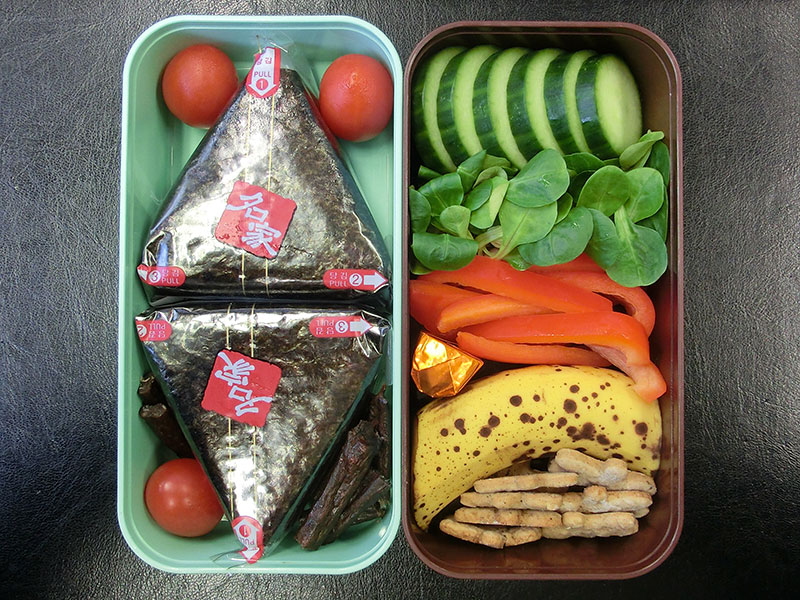 Bento Box gefüllt mit Onigiri, Galloway Sticks, Tomaten, Gurke, Paprika, Schokolade, Banane, Kekse