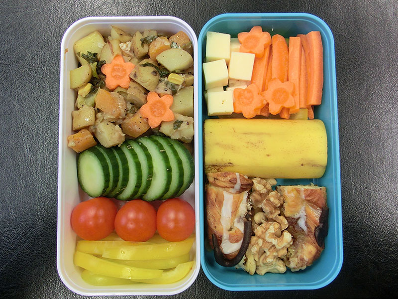 Bento Box gefüllt mit Tomaten, Gurke, Paprika, Kartoffelpfanne, Kase, Karotten, Bananen, Kaneelstang, Nüsse